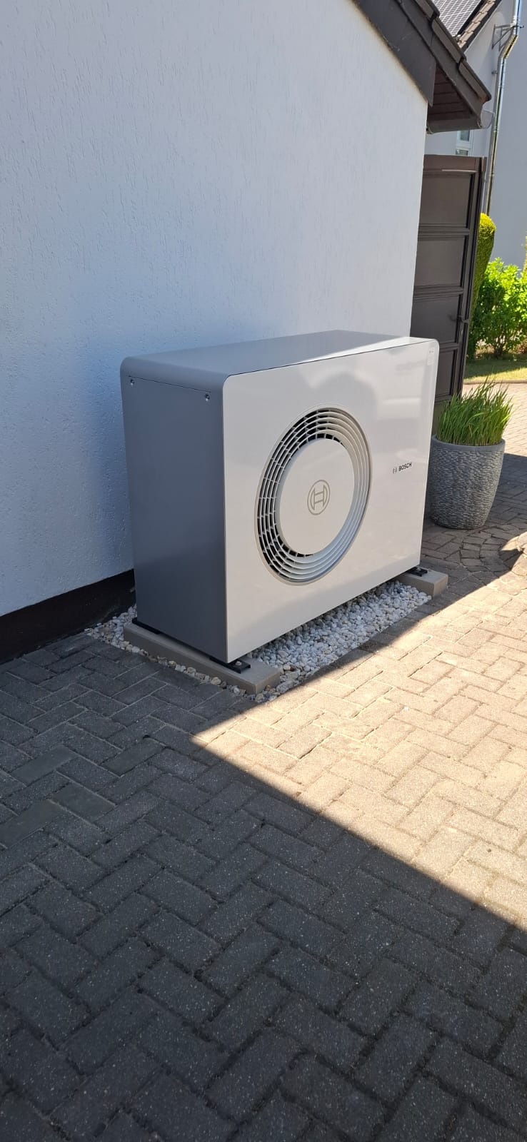 Außenstehende Bosch-Wärmepumpe an Hauswand auf Kiesbett mit Pflastersteinen im Vordergrund bei Tageslicht.